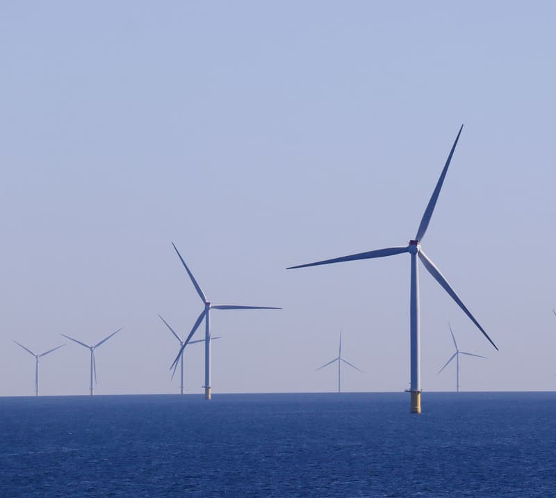 Row of offshore wind turbines at sea, blades turning against a pale sky