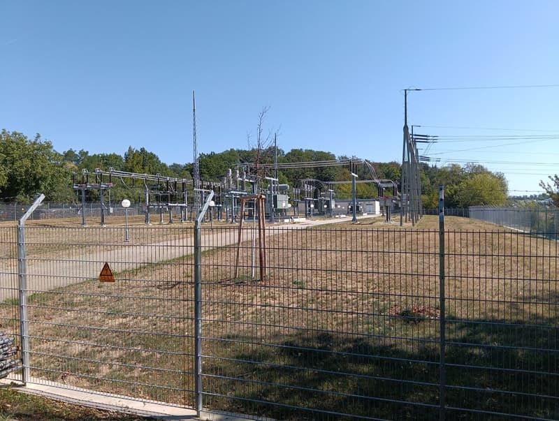 An electrical substation switchyard with busbars, circuit breakers and transmission towers behind a security fence